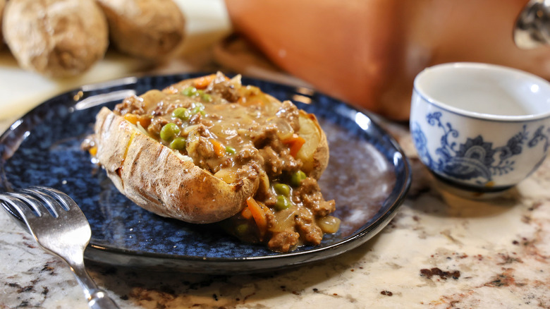 jacket potato filled with beef and vegetables in gravy on a blue plate with a fork and salt cellar beside it
