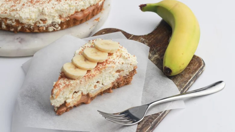 Slice of banoffee pie on cutting board with a whole unpeeled banana on the side