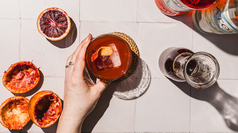 Hand holding a glass with blood orange Screwdriver cocktail alongside empty blood orange peels and bottles