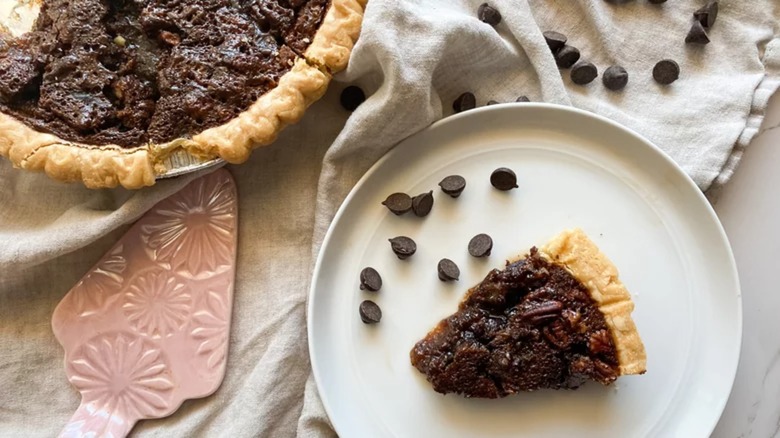 Slice of chocolate pecan pie next to a whole pie and chocolate chips