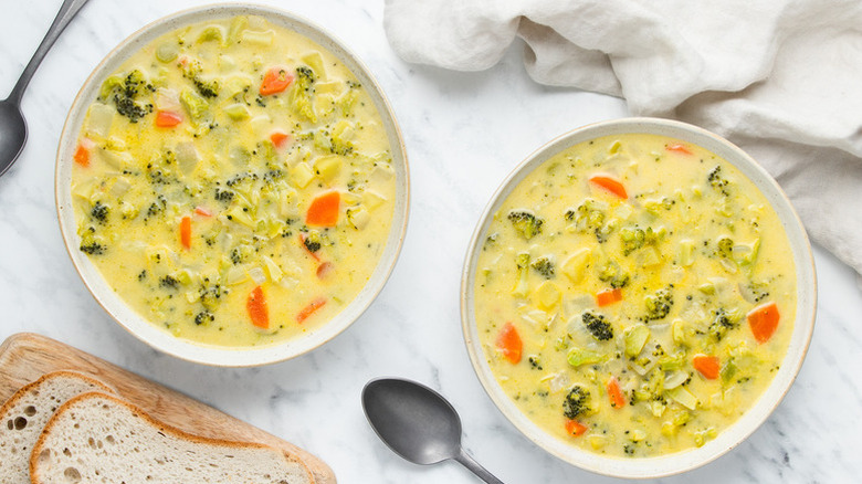 Two bowls of broccoli cheddar soup next to spoons and bread slices