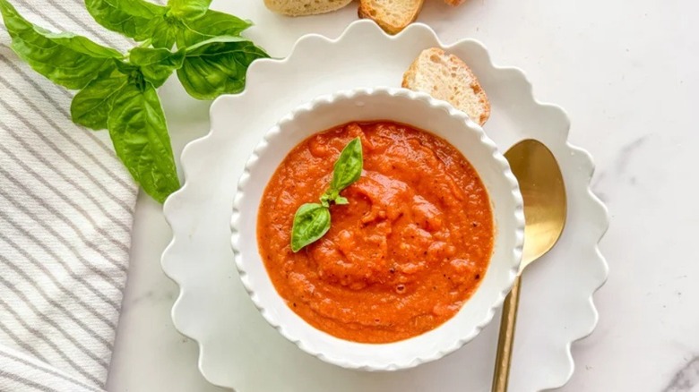 Bowl of tomato soup garnished with basil next to spoon and bread