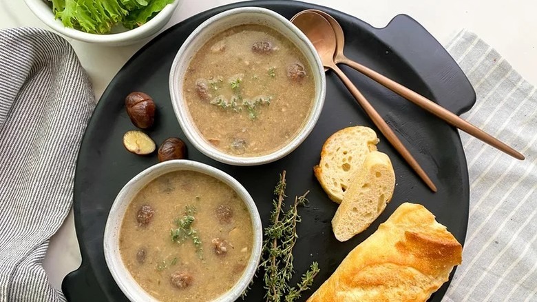 Two bowls of soup on platter with chestnuts, baguette, thyme, and two spoons