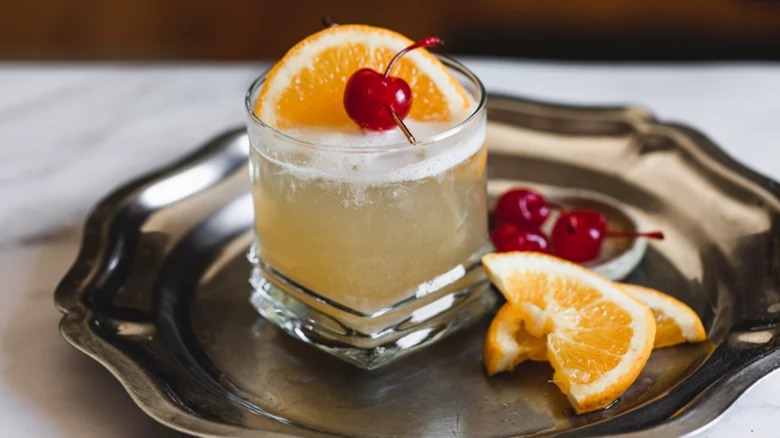 Frothy whiskey sour on silver tray with orange slice and maraschino cherry garnishes