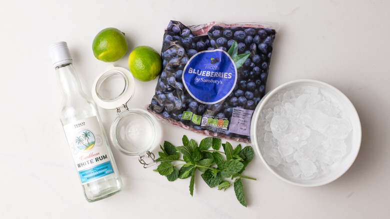 bag of frozen blueberries, two limes, bottle of white rum, mint leaves, bowl of ice, and jar of simple syrup laid out