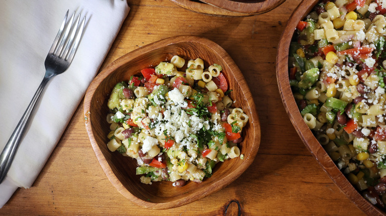 an overhead view of a wooden bowl of cowboy caviar pasta salad with a fork and napkin as well as a larger serving bowl beside it
