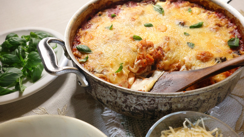 wooden spatula in pan of gnocchi lasagna garnished with basil leaves beside plate of fresh basil