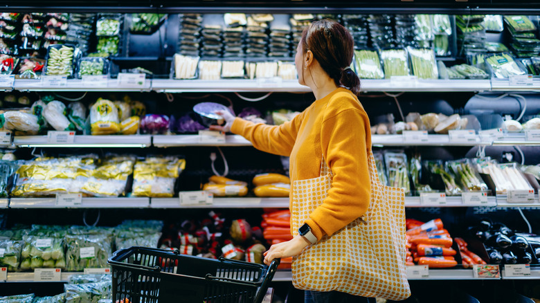 A person in a yellow top with a bag on their arm pushing a grocery cart and selecting produce from the shelves at a grocery store