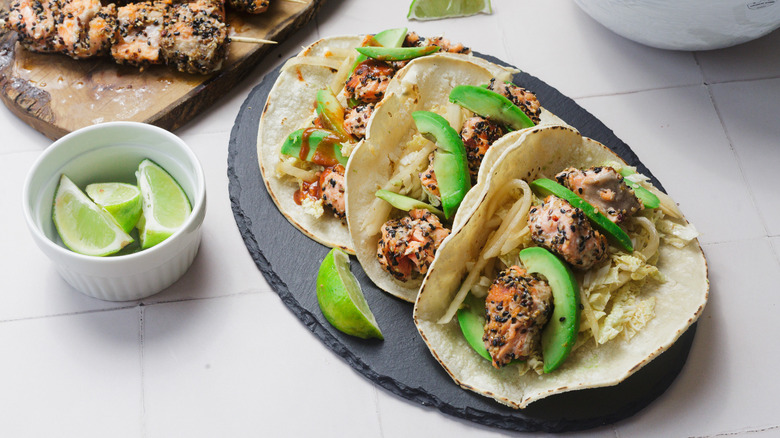 three salmon tacos with lime wedges and more salmon skewers in the background