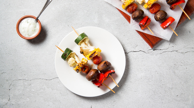 two cooked veggie skewers served on white plate next to more veggie skewers and yogurt sauce
