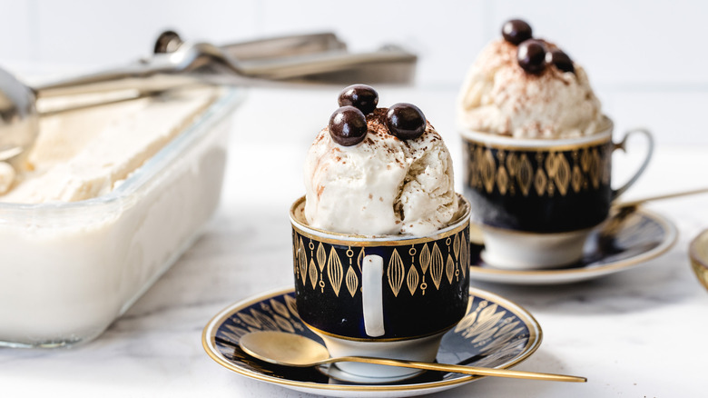 Two coffee cups with ice cream and container with ice cream scoop in background