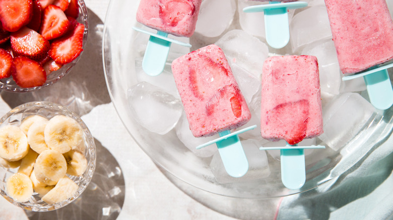 fruit popsicles arranged over ice on table surrounded by sliced strawberries and bananas