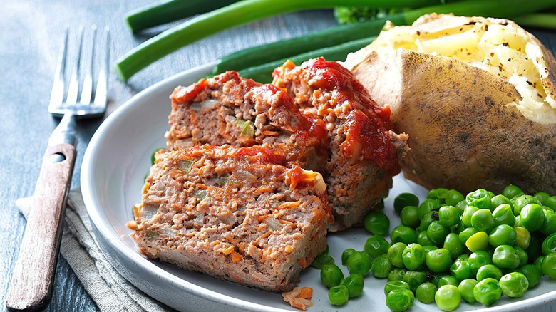 Three slices of meatloaf on plate next to baked potato and peas