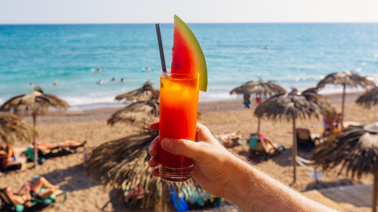 someone holding a watermelon cocktail with the beach in the background