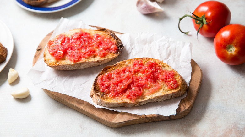 pan con tomate on lined wooden board next to garlic cloves and whole tomatoes