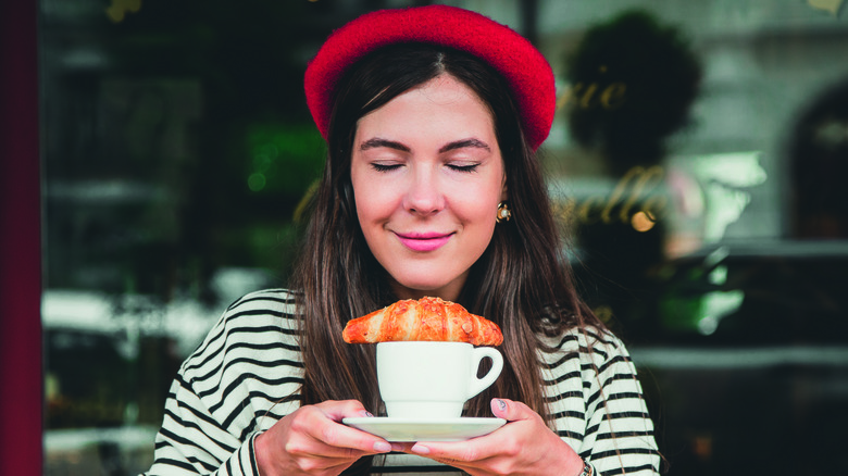 Woman in red beret and breton shirt with eyes closed holding coffee with croissant