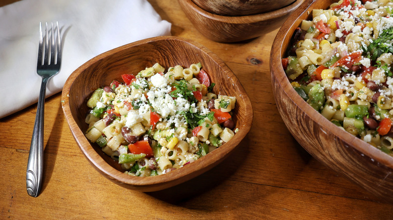 a wooden bowl of cowboy caviar pasta salad next to a wooden werving bowl of the same as well as a fork and napkin on a wooden table