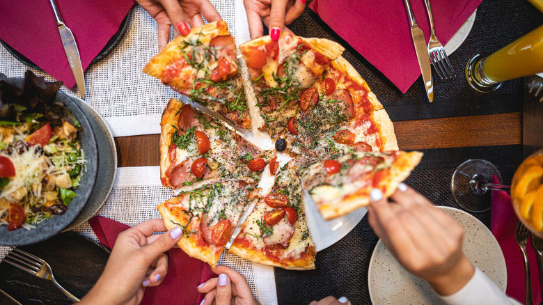 Top-down view of hands taking slices from a pizza on table with cutlery and salad