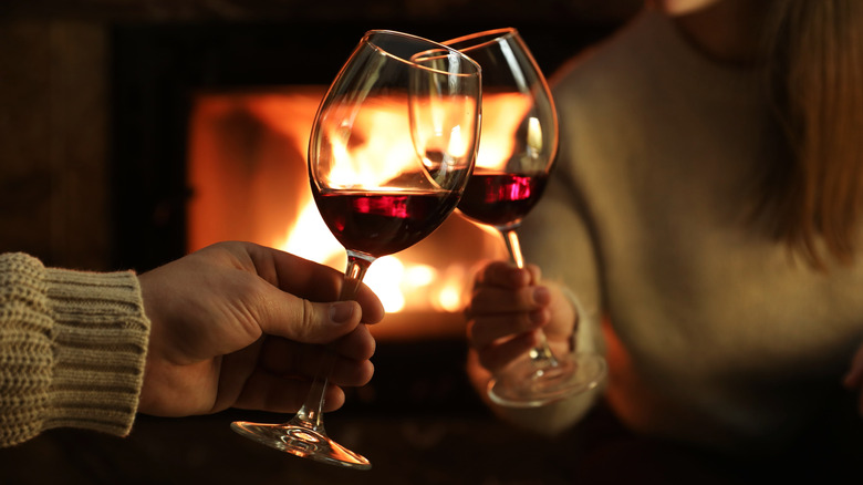 Two people clinking red wine glasses by fireplace