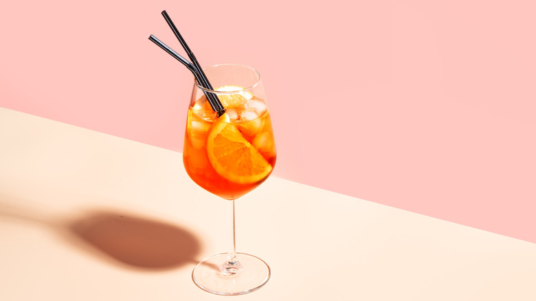 Aperol spritz in a wine glass with black straws and an orange slice on pink background
