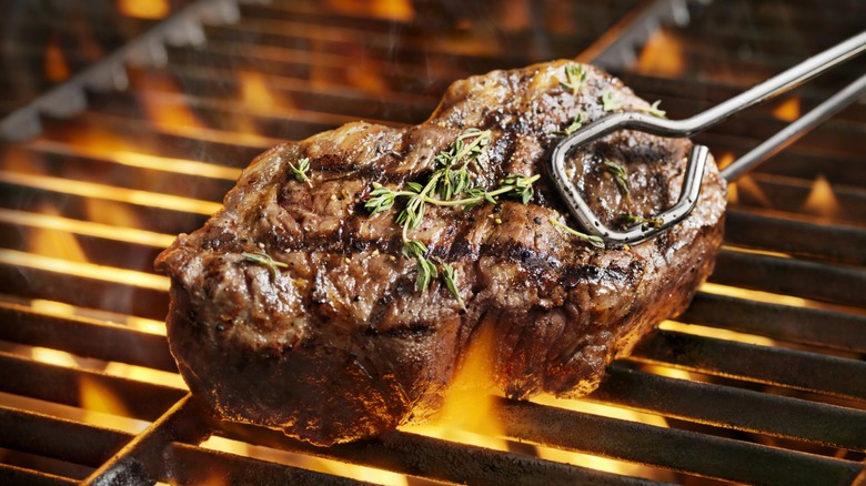 an herbed steak, covered in grill marks, held by tongs above a flaming grill