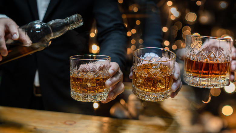 Multiple hands holding out rocks glasses full of bourbon while one person pours the bourbon from the bottle