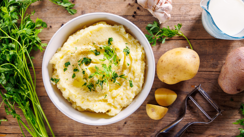 Bowl of mashed potatoes garnished with fresh herbs on a wooden table surrounded by herbs, potatoes, garlic, and cream