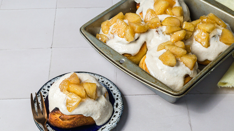 One cinnamon bun on a small plate in front of tray of cinnamon buns