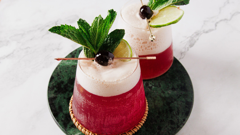 Two pink frothy cocktails in glasses, garnished with fresh mint, lime, and cherries