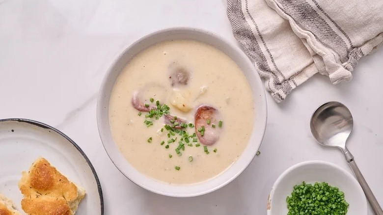 Bowl of potato and sausage soup topped with chives, surrounded by napkin, spoon, bread, and bowl of chives