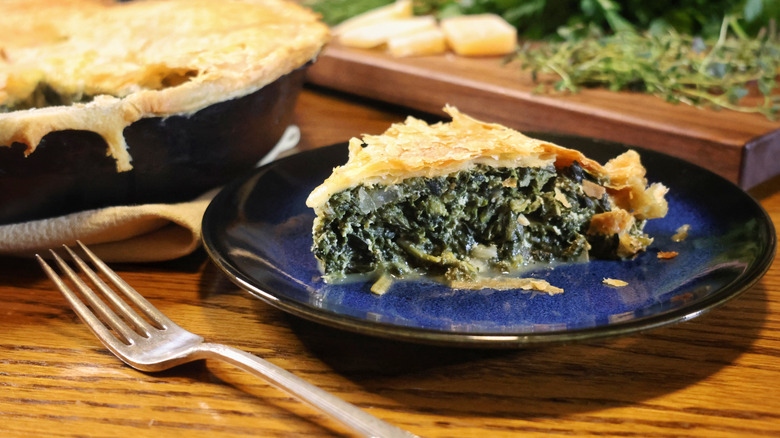 slice of spinach pie on plate beside pie and cutting board with cheese and herbs