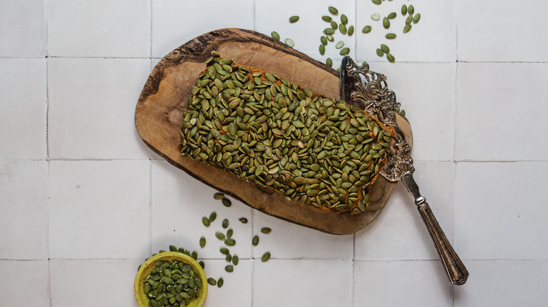 A slice of pumpkin bread on small plate next to sliced pumpkin bread on a cutting board next to bowl of pumpkin seeds
