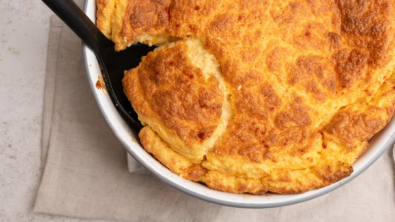 A big spoon dug into a baked cheesy corn souffle sitting on top of a cloth