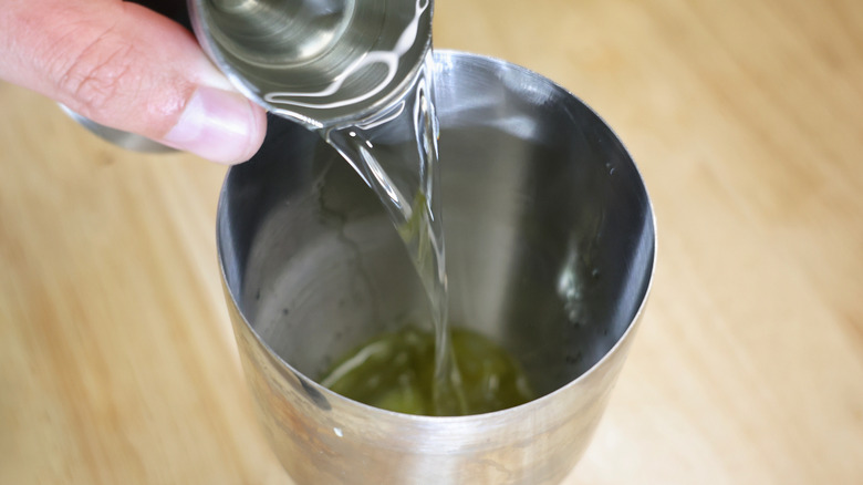 hand pouring clear liquid from shot glass into cocktail shaker