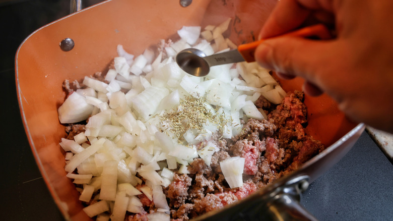 hand dumping measuring spoon of dried rosemary into pan of onions and ground beef