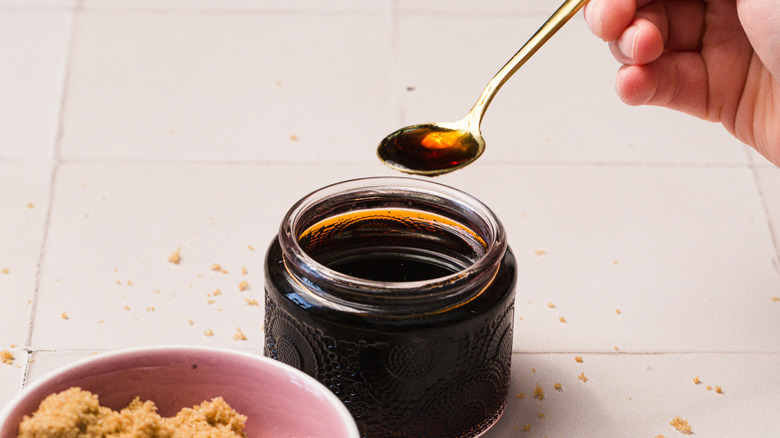 A hand lifting a spoon of sugar over a jar of syrup