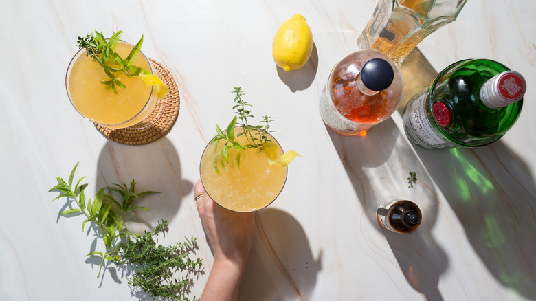 Hand holding a yellow cocktail out of two, standing next to lemon verbena and lemon and liqueur bottles