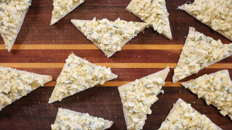 triangles of bread with egg salad on them on a wooden cutting board