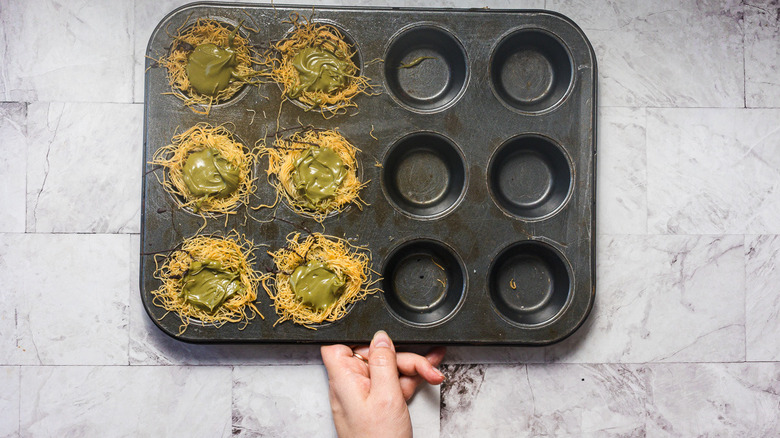 Holding muffin tin with kataifi noodle nests filled with pistachio cream