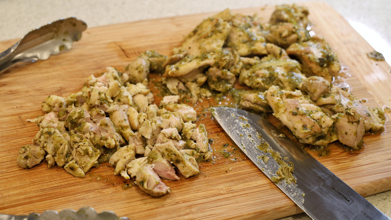 chopped salsa verde chicken thighs on a wooden cutting board with a chef's knife and tongs