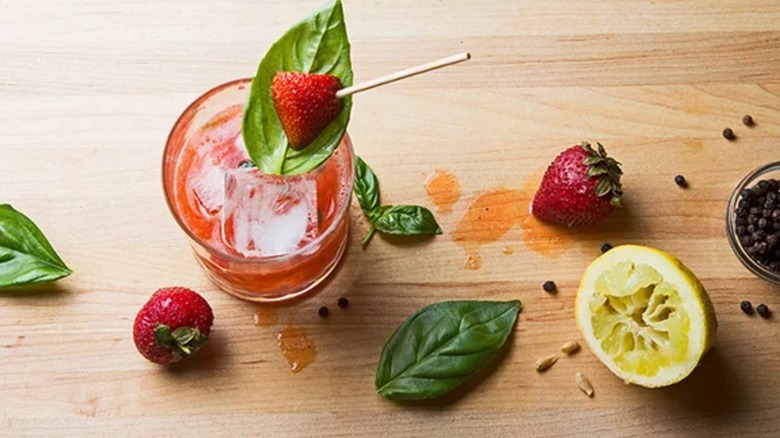 Strawberry peppercorn smash in a glass with ice, basil, strawberries, limes, and peppercorns