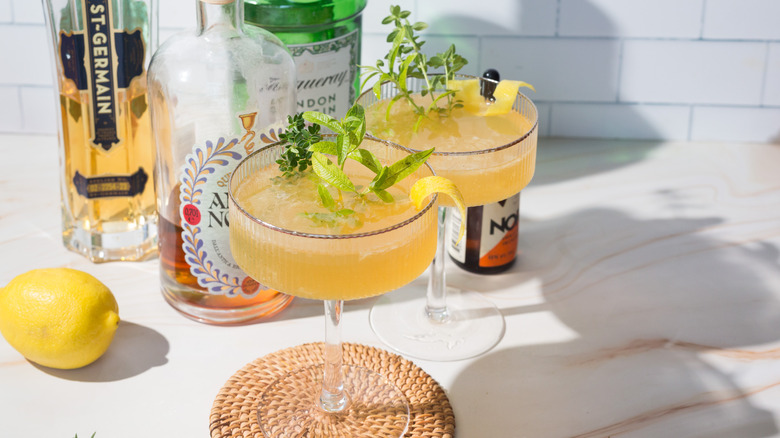 Two yellow cocktails next to lemon verbena and lemon and liqueur bottles