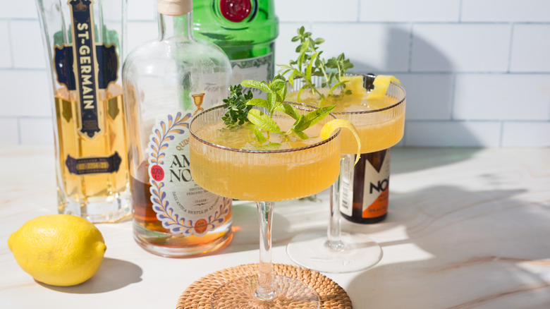 Two yellow cocktails next to lemon verbena and lemon and liqueur bottles