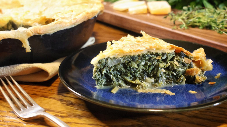 slice of spinach pie on plate with fork, cast iron pan, and cutting board beside it